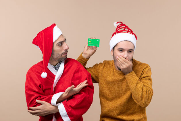 米色前视图两个家伙一个穿着圣诞外套 另一个拿着信用卡在米色的孤立背景上闭着鼻子伙计们外套成人