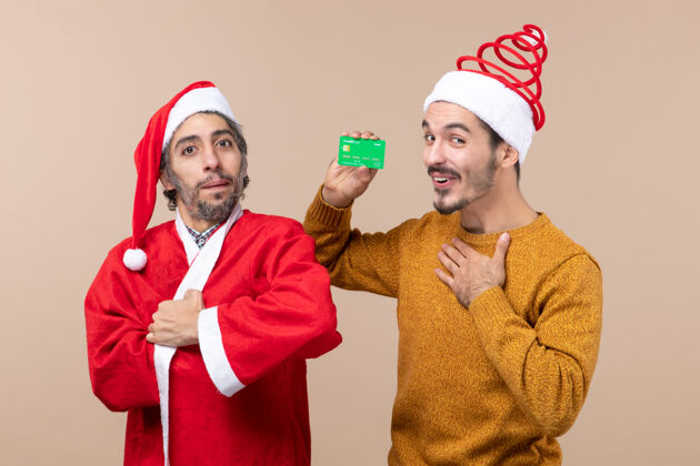 圣诞老人正面图两个朋友 一个穿着圣诞外套 另一个手放在胸前 背景是米色的朋友前面男