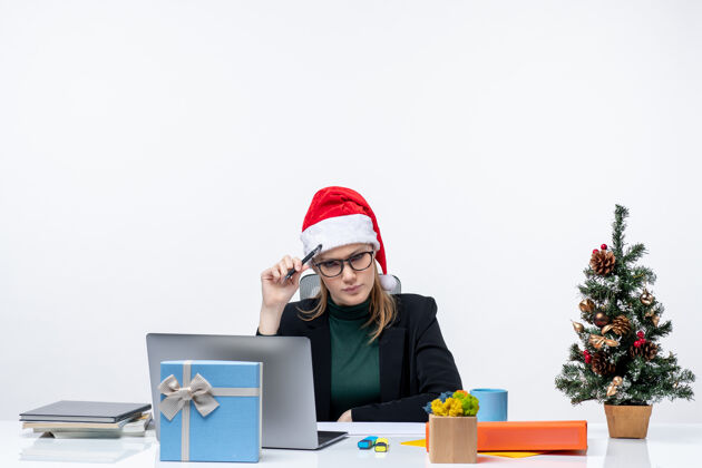 公司新年的气氛与犹豫不决的金发女人坐在一张桌子上 圣诞树和礼物的白色背景圣诞老人帽子金发女人礼物女人