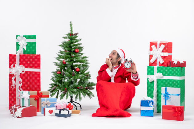 礼物惊喜情绪化的年轻人庆祝新年或圣诞节假期坐在地上 拿着礼物和装饰圣诞树附近的白色背景时钟男人坐着圣诞树