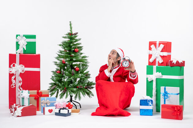 时钟震惊的年轻人庆祝新年或圣诞节假期坐在地上 拿着礼物和装饰圣诞树附近的时钟礼物年轻人年轻人