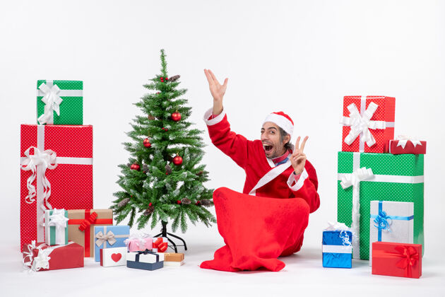 礼物滑稽的年轻人打扮成圣诞老人 拿着礼物和装饰好的圣诞树坐在地上 指着上面 在白色背景上做着胜利的手势圣诞地上成年人