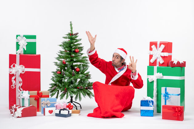 眼镜滑稽的年轻人打扮成圣诞老人 拿着礼物和装饰好的圣诞树坐在地上指指点点 在白色背景上做眼镜手势年轻人坐着圣诞