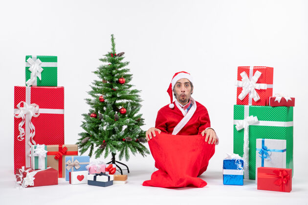 震惊震惊的年轻人打扮成圣诞老人与礼物和装饰圣诞树坐在地上寻找白色背景的东西年轻人礼物大人