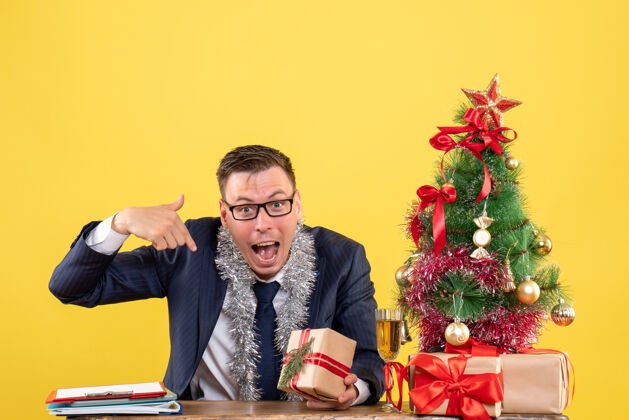 树年轻人指着自己坐在圣诞树旁的桌子 在黄色的礼物男人圣诞桌子
