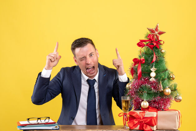 指着一个年轻人站在圣诞树旁边的桌子旁 用手指着舌头 用黄色的礼物礼物手指桌子