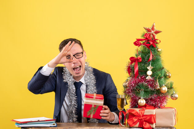 黄色在圣诞树旁的桌子旁 一个年轻人在眼前做着“ok”的手势 并在黄色的纸上做着礼物男性制作礼物