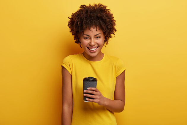 女性照片中快乐的非洲裔美国妇女手持外卖咖啡 享受芳香饮料 有着牙齿般的微笑 洁白的牙齿 穿着休闲的黄色t恤人类发型杯子