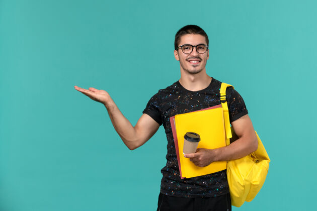 背包身穿深色t恤黄色背包的男学生正面图 蓝色墙上挂着微笑的咖啡 手里拿着不同的文件书持有黑暗