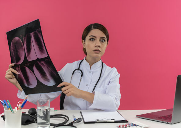 电脑惊讶的年轻女医生穿着医用长袍带听诊器坐在办公桌上用医疗工具拿着电脑拿着x光片有复印空间工作穿着听诊器