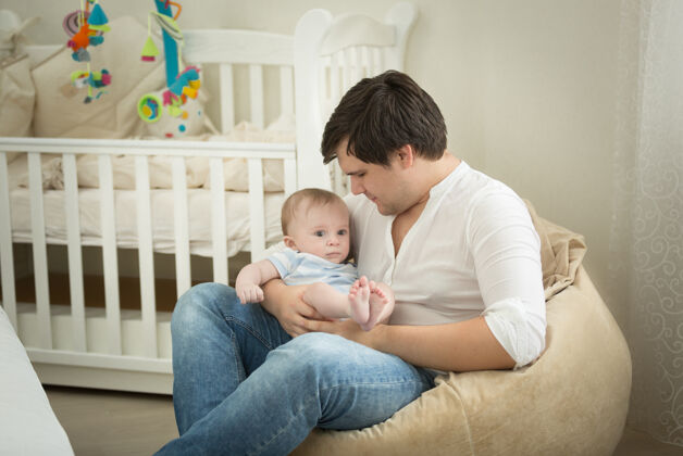 休闲年轻的父亲坐在卧室里 双手抱着他的小儿子婴儿亲情床