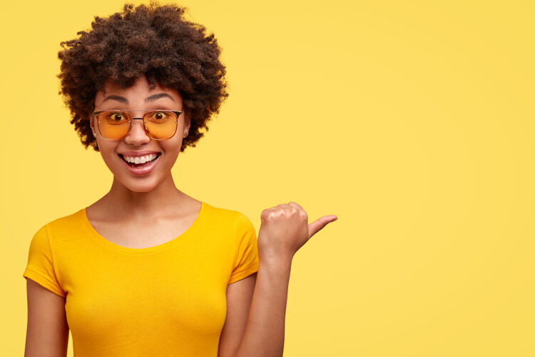 高兴快乐的黑皮肤女人 非洲式发型 拇指指向一边 对大减价很满意 戴着太阳镜和休闲t恤自信个人外观