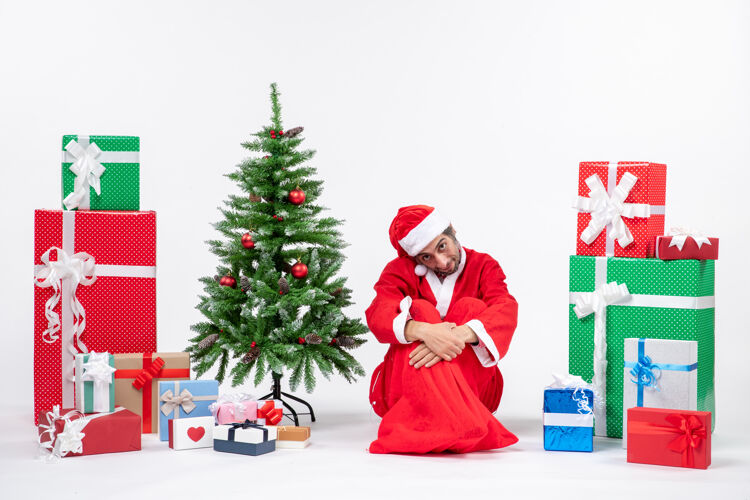 礼物悲伤不满足的年轻人打扮成圣诞老人 拿着礼物和装饰好的圣诞树坐在白色背景的地上年轻人圣诞老人圣诞老人