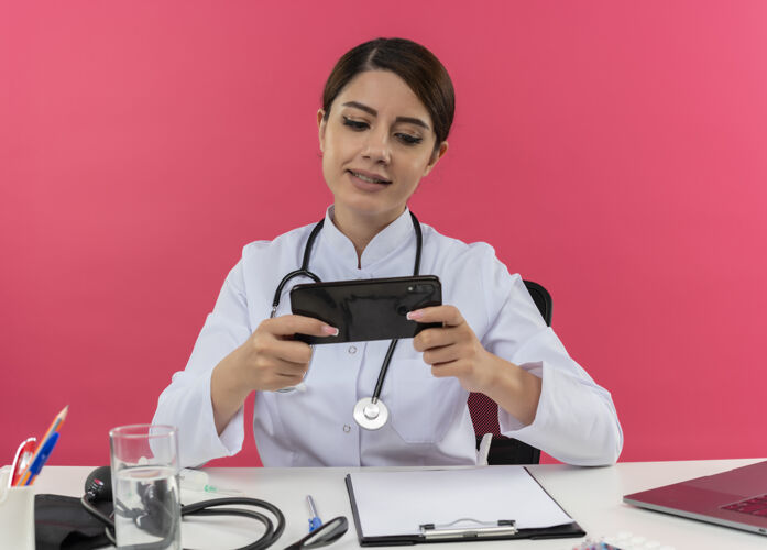 医疗高兴的年轻女医生穿着医用长袍 听诊器 坐在办公桌上 用电脑和医疗工具 在手机上玩游戏 还有复印空间玩电话坐