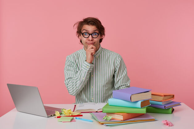 笔记本一头野性头发的积极的年轻男性坐在工作桌旁 拿着书和笔记本电脑 穿着衬衫和眼镜摆姿势 手托下巴 扬眉吐气眼镜宽学习