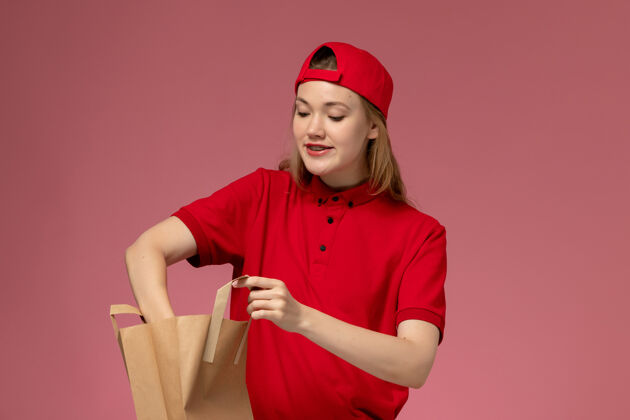 东西正面图身穿红色制服和披风的年轻女快递员拿着快递食品包 在粉色墙上从里面拿东西肖像制服人