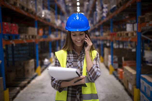 手册美丽的女仓库工人在大型仓储配送中心用手机交谈的画像智能电话职业安全