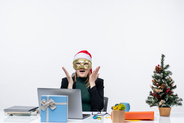 惊讶圣诞节的心情与惊喜兴奋的年轻女子戴着圣诞老人的帽子 戴着面具坐在一张白色背景的桌子笔记本面具女人
