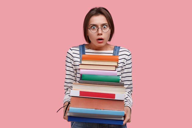 Omg受惊的女学生背着一堆手册 戴着眼镜和条纹毛衣 愤怒地看着镜头震惊反应恐惧