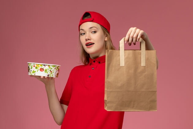 工作正面图：身穿红色制服和披风的年轻女快递员拿着快递食品包和碗放在粉红色的服务台上工作制服快递前面红色包裹