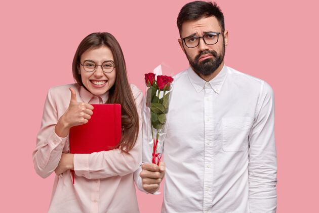 女性美女的照片展示了赞同的姿态 竖起大拇指 拿着红色的记事本 困惑的没刮胡子的男人拿着一束玫瑰花玫瑰享受眼镜
