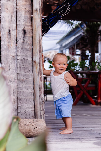 漂亮穿着蓝色短裤和白色衬衫的九个月大的快乐孩子的户外肖像站在木墙上微笑着欢快泰国蹒跚学步
