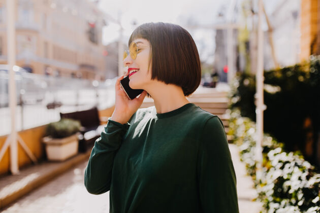 户外迷人的短发女人在打电话穿着绿色毛衣的漂亮黑发女孩在街上给别人打电话享受手机快乐