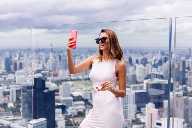城市年轻快乐微笑的高加索女旅客 穿着合身的衣服 戴着墨镜站在曼谷的高楼上 手里拿着电话站立中心城市景观