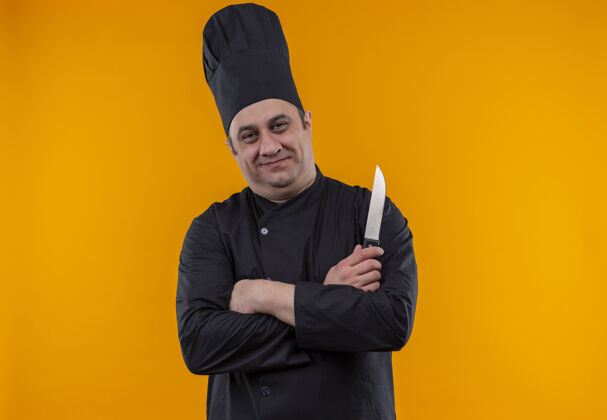 手喜出望外的中年男厨师身着厨师制服双手持刀交叉在黄墙上刀中年制服