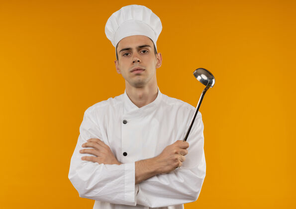 酷年轻男酷穿厨师制服交叉手拿勺子在孤立的黄色墙上手穿抱