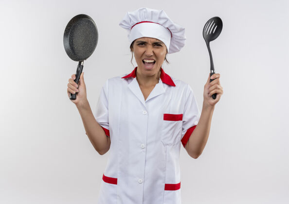 平底锅愤怒的年轻女厨师穿着厨师制服 拿着煎锅和抹刀 站在隔离的白色墙壁上 留着复印空间愤怒穿年轻