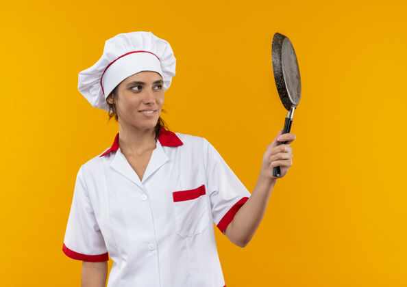 平底锅年轻的女厨师面带微笑 身穿厨师制服 手拿煎锅 看着有复印空间的煎锅穿着厨师女