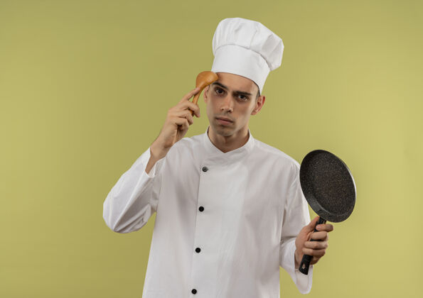 厨师困惑的年轻男厨师穿着厨师制服拿着煎锅把勺子放在额头上孤立的绿色墙壁上拿看穿