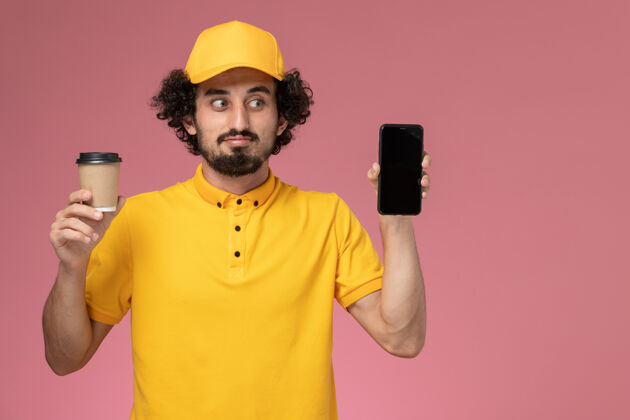 咖啡正面图：身穿黄色制服 披风的男性快递员 在粉色墙上拿着送货咖啡杯和电话帅哥男信使制服