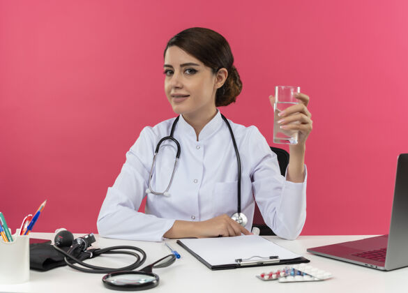 穿高兴的年轻女医生穿着医用长袍 带听诊器 坐在办公桌旁 拿着医疗工具 拿着一杯水 留着复印空间长袍坐水