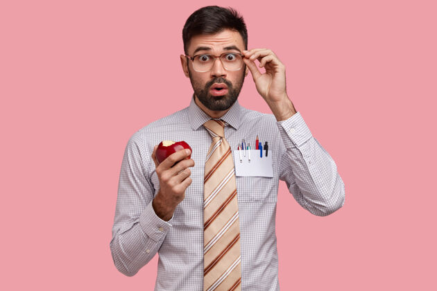 知识震惊的商人穿着正式的衬衫和领带 吃着美味的苹果 目不转睛地看着眼镜没有刮胡子铅笔眼镜