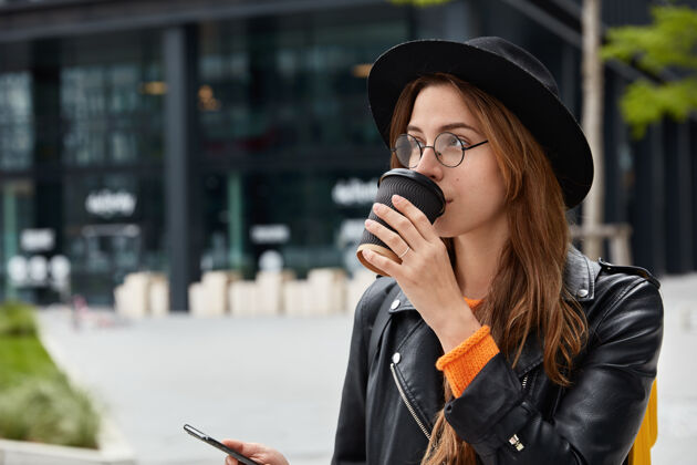 持有沉思的少女侧着镜头漫步城市后喝咖啡休息 手持智能手机设备 查看邮箱 聚焦远方咖啡科技服装