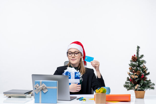 办公室戴着圣诞老人帽戴着眼镜的忙碌的年轻女子坐在桌子旁 手里拿着圣诞礼物和白色背景的银行卡公司笔记本电脑年轻人