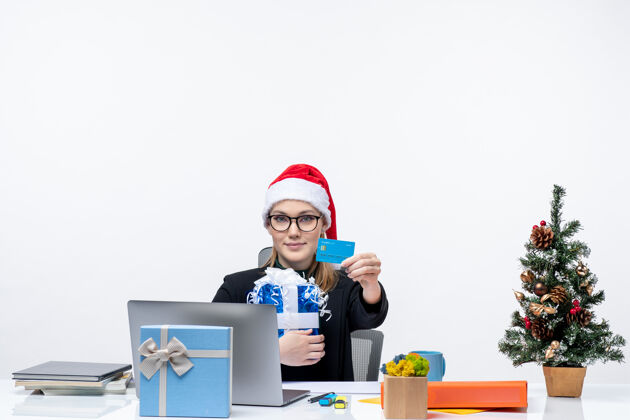 人戴着圣诞老人帽 戴着眼镜的快乐年轻女子坐在桌子旁 手里拿着圣诞礼物 手里拿着白色背景的银行卡桌子银行圣诞老人