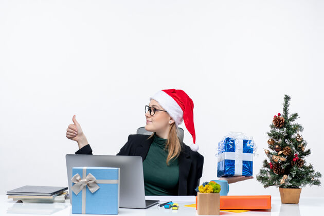 圣诞老人带着圣诞老人帽子戴着眼镜的年轻女子坐在桌旁展示圣诞礼物 指着右边的东西做着“ok”的手势帽子年轻圣诞老人