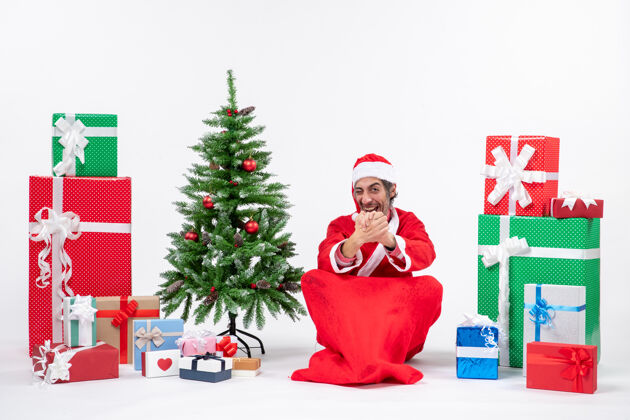 地面惊讶的年轻人打扮成圣诞老人和礼物装饰圣诞树坐在地上的白色背景年轻人圣诞老人成人