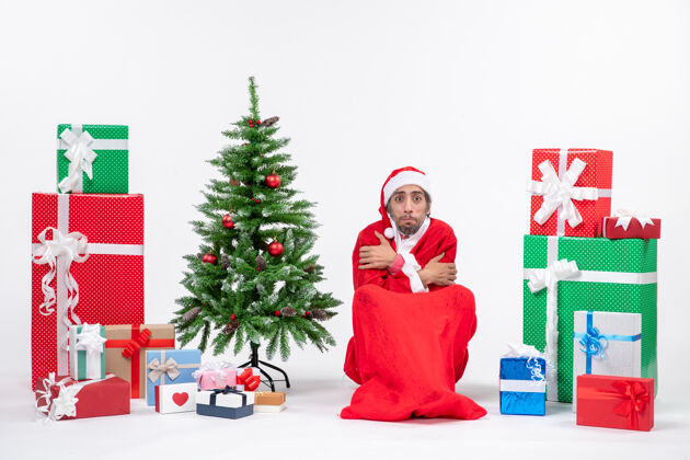 圣诞树冻僵的年轻人打扮成圣诞老人 拿着礼物和装饰好的圣诞树坐在白色背景的地上礼物年轻人礼物
