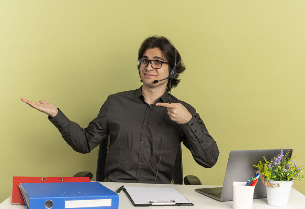 空间一个戴着耳机戴着眼镜的年轻人坐在办公桌旁 手里拿着办公工具 手里空空拿着笔记本电脑点工人笔记本电脑
