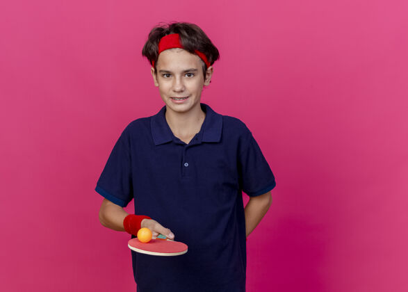 高兴高兴的年轻帅气的运动男孩戴着头带和腕带 拿着乒乓球拍和球 保持手后面孤立青少年背深红色