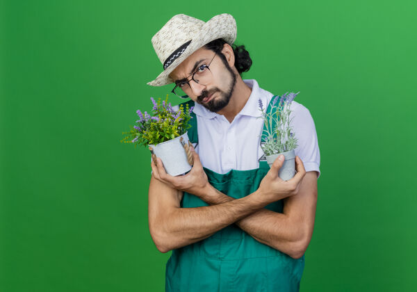 胡须年轻的留着胡子的园丁 穿着连体衣 戴着帽子 手里拿着盆栽植物 严肃的脸皱着眉头 不高兴帽子绿色植物