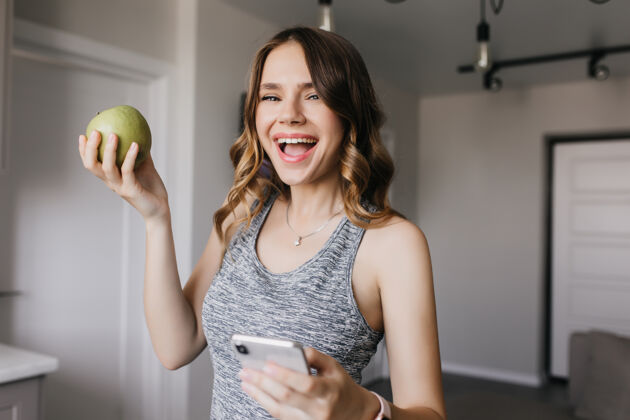 饮食运动型女孩在与水果合影时表达快乐室内拍摄轻松卷曲的女人用苹果和智能手机金发配件智能手机