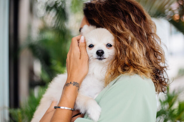 幸福欧洲棕褐色卷曲女人的户外肖像抱着快乐的宠物狗波美拉尼亚斯皮茨纯种夏天在一起