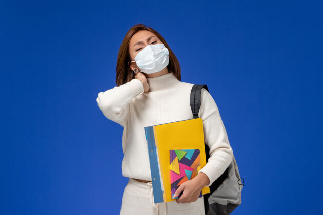 肖像正面图身穿白色运动衫的年轻女学生戴着带包的面具 蓝色墙上有脖子疼的抄写本教训成人文案