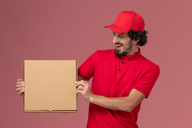 成人正面图：身穿红色衬衫和披风的男性快递员手持浅粉色墙上的送货食品箱男性快递披萨人