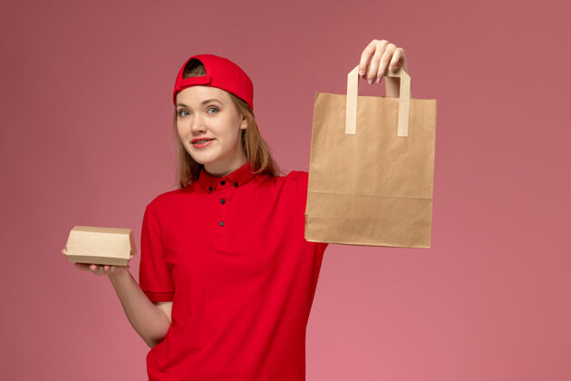 微笑正面图：身穿红色制服 披风的年轻女快递员在粉色墙上拿着快递食品包服务工作专业